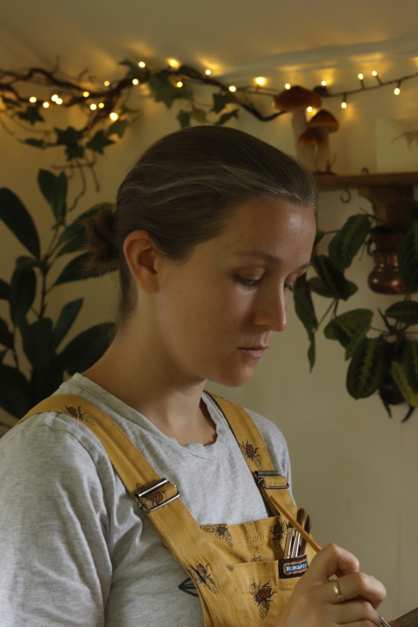 A side profile photograph of artist Annie Drew working in her studio, wearing yellow dungarees patterned with bees and holding a fine paintbrush against a backdrop of warm fairy lights and green houseplants.